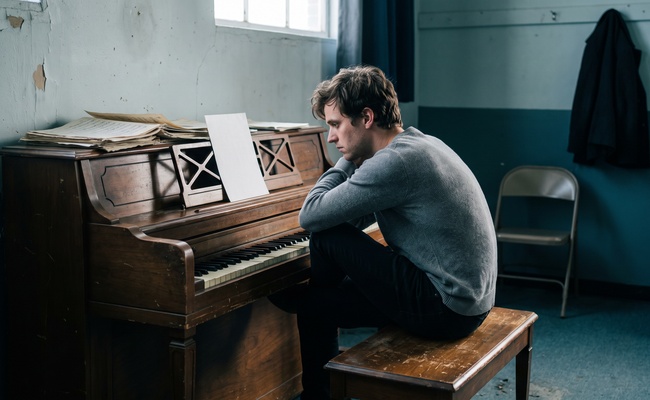 Man seated sideways on a piano bench reading a single page propped on the music stand.