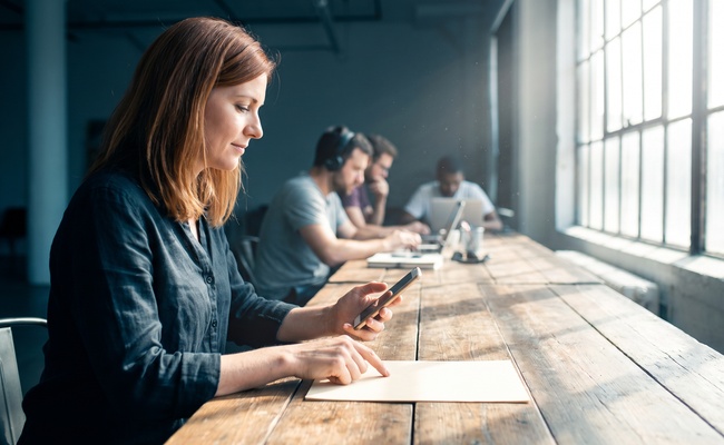 Woman at a long shared work table reading her phone carefully with one finger resting on a sheet of paper.