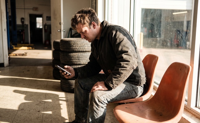 A young man sitting in a waiting area checks his phone carefully to confirm he captured a collection message correctly.