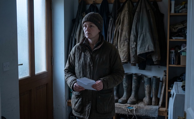 A woman still wearing her coat stands in a mudroom at dawn holding an opened envelope she was not expecting.