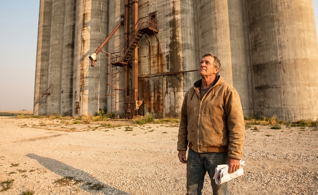 A man in a work jacket holds folded paperwork at the base of a grain elevator while he weighs what he actually signed.