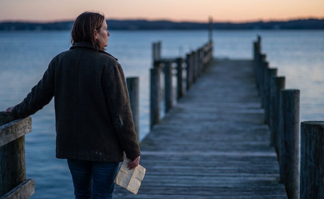 A woman rests one hand on a weathered pier rail at dusk holding a folded letter threatening to garnish a spouse's wages.