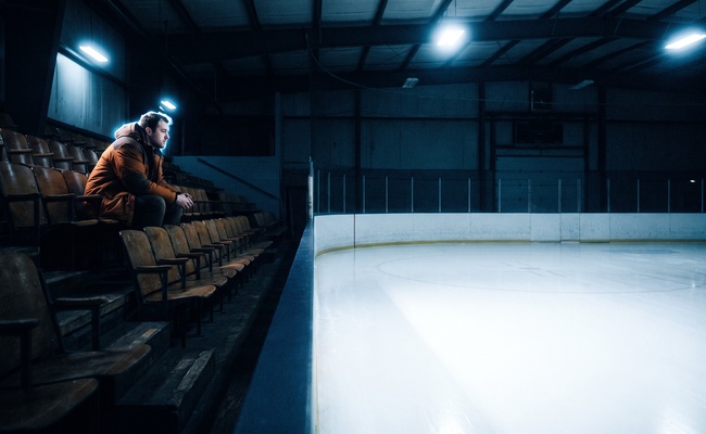 A man sits alone high in empty rink bleachers in cold light, the waiting that follows filing a claim and precedes a hearing date.