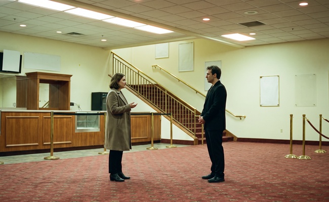 Two people stand a measured distance apart in an empty lobby under full house lights, the plain businesslike exchange most exemption hearings actually resemble.