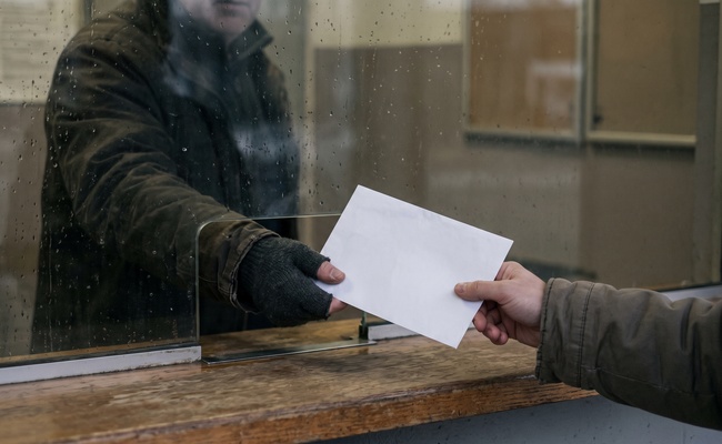 A plain envelope passed across a worn ledge at a parcel window, standing for the notice a bank is required to send after it freezes an account.