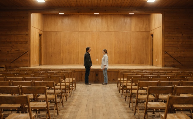Two people talking on a low stage in an empty hall of wooden chairs, conveying how ordinary and businesslike a levy exemption hearing is.