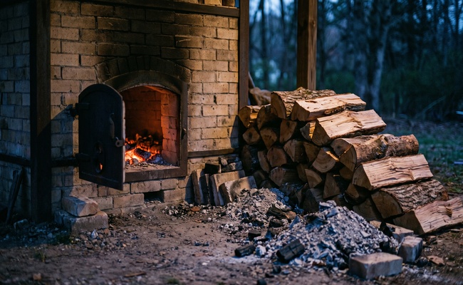 An open kiln door glowing with coals beside a stacked cord of split pine, heat committed before the door is closed, the risk of paying to settle a debt before judgment too early.