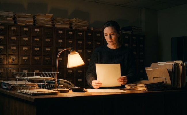 Financial aid office clerk holding a blank sheet at her counter after closing, before she learns she must remove identity theft accounts from her own file