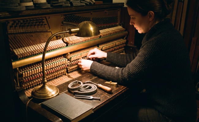 Piano tuner adjusting dampers beside a closed exercise book on the bench