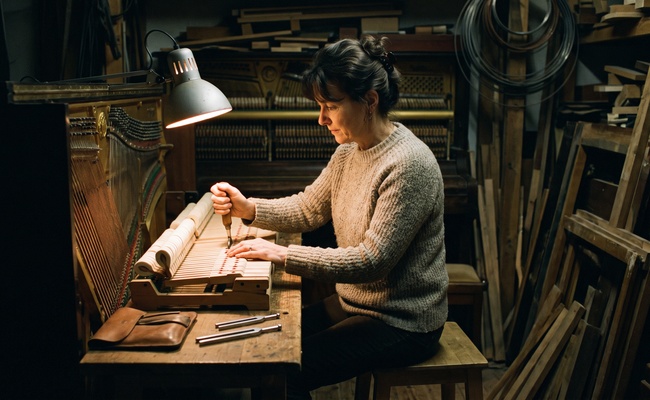 A piano tuner comparing two tuning forks at a workshop bench in evening light