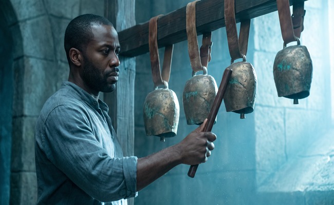 A person draws a wooden beater across four graduated bronze bells hanging in a row.